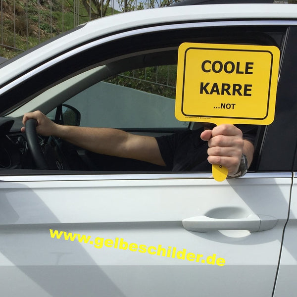 Autofahrer hält gelbes Schild mit Textaufschrift "Coole Karre...NOT" aus dem Fenster 
