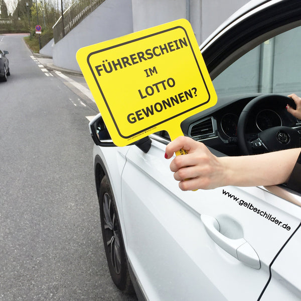 Autofahrerin hält gelbes Schild mit Textaufschrift "Führerschein im Lotto gewonnen" aus dem Fenster 