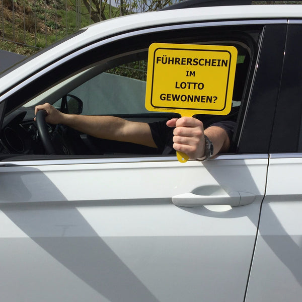 Autofahrer hält gelbes Schild mit Text "Führerschein im Lotto gewonnen?" aus dem Auto