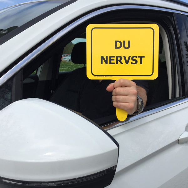Autofahrer hält gelbes Schild mit Textaufschrift "Du nervst" aus Fenster von weißem Auto