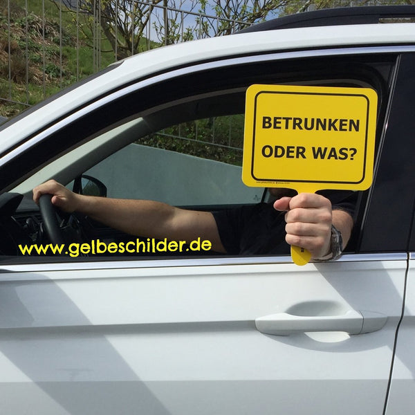 Autofahrer hält gelbes Schild mit Textaufschrift "Betrunken oder was" aus dem Fenster