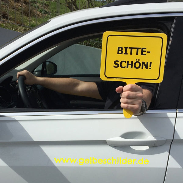 Autofahrer hält gelbes Schild mit Textaufschrift "Bitteschön!" aus dem Fenster 