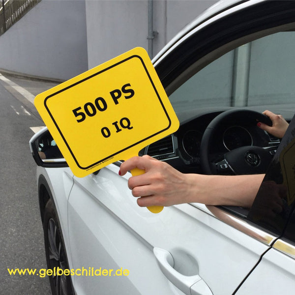 Autofahrerin hält gelbes Schild mit Textaufschrift "500 PS - 0 IQ" aus dem Fenster 