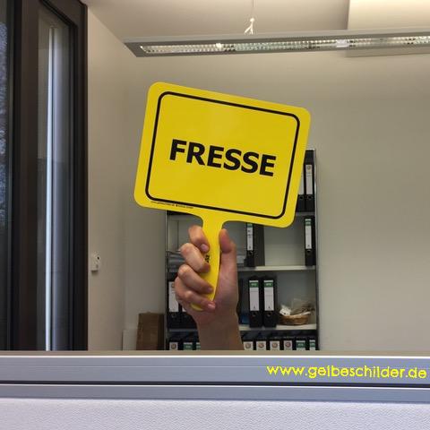 Kollege hält gelbes Schild mit Textaufschrift "Fresse" im Büro hoch