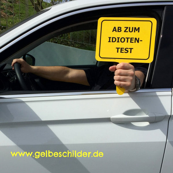 Autofahrer hält gelbes Schild mit Textaufschrift "Ab zum Idiotentest" aus dem Fenster 